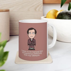 May include: White ceramic mug with a brown background featuring a cartoon illustration of a man in a red and black plaid robe. The text on the mug reads "I seem to be having this tremendous difficulty with my lifestyle."