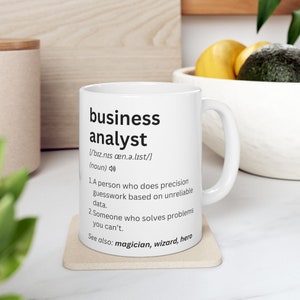 Peut inclure: Mug en céramique blanche avec l'inscription "business analyst" en noir, accompagnée d'une définition. Le mug est posé sur un dessous de verre beige. La définition comprend la prononciation, une description et des termes associés.