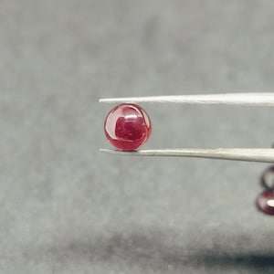 May include: A round, red gemstone held in tweezers. The gemstone is smooth and shiny.