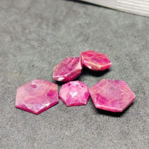 May include: Five pink ruby gemstones, cut into hexagonal shapes, are arranged on a gray surface. The gemstones are faceted and have a shiny, polished finish.