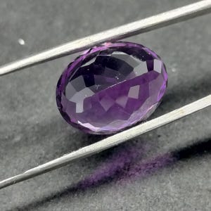May include: A purple oval-shaped amethyst gemstone with a faceted cut. The gemstone is held in place by a pair of tweezers.