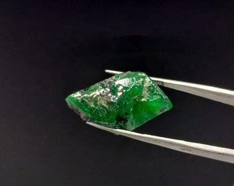 May include: A vibrant green emerald gemstone with a faceted cut, held by tweezers against a black background.