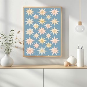 May include: A rectangular wall art print in a light wooden frame featuring a repeating pattern of multi-pointed stars in shades of pale yellow, cream, and soft pink against a solid dusty blue background, displayed in a vertical orientation.