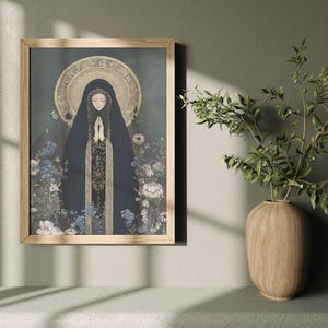 May include: A framed art print featuring a religious figure in a dark blue cloak with hands clasped in prayer. The artwork has a gold halo and is surrounded by blue and white flowers. The frame is made of light-colored wood.