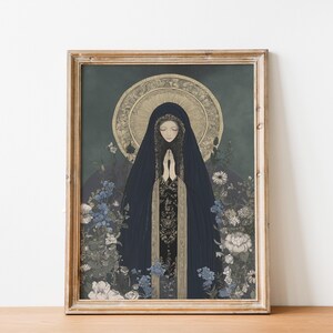 May include: Framed art print featuring a woman in a dark blue hooded cloak with hands clasped in prayer. The artwork has a gold halo and is surrounded by blue and white flowers. The frame is made of light brown wood.