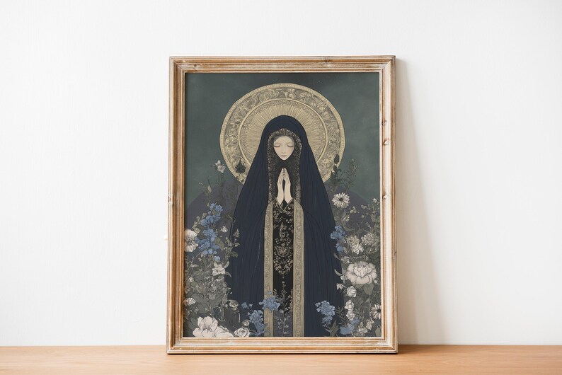May include: Framed art print featuring a woman in a dark blue hooded cloak with hands clasped in prayer. The artwork has a gold halo and is surrounded by blue and white flowers. The frame is made of light brown wood.