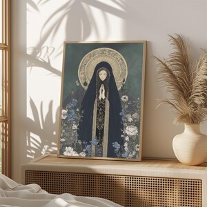 May include: A framed art print featuring a religious figure in a dark blue veil with hands clasped in prayer. The artwork has a gold halo and is surrounded by blue and white flowers. The print is displayed on a wooden surface.