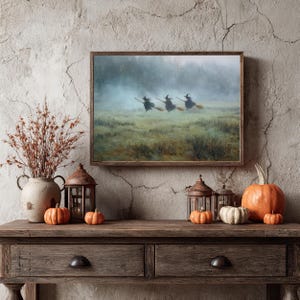 May include: A framed print depicts three witches on broomsticks flying over a misty field. The artwork is displayed above a wooden console table decorated with pumpkins, lanterns, and a vase of dried branches, creating a Halloween theme.