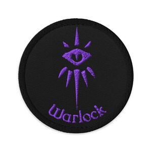 May include: Round black patch with a purple eye and stylized rays design. The word "Warlock" is embroidered in purple script below the eye symbol. The patch has a black border.
