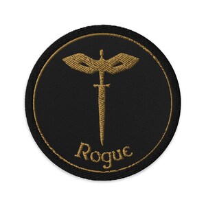 May include: Embroidered patch with a black background and a gold-colored design. The design features a stylized sword with a wing-like shape above it, and the word "Rogue" in a script font below. The patch has a gold border.