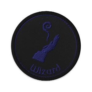 May include: A round, black embroidered patch with a purple wizard-themed design. The design features a hand with a swirling energy effect and the word "Wizard" in purple script. The patch has a purple border.