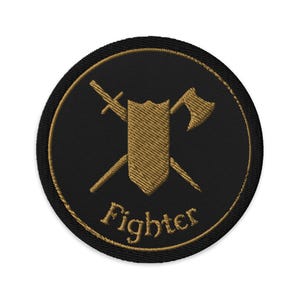 Puede incluir: Parche bordado con fondo negro y borde dorado. El parche presenta un diseño dorado de escudo, espada y hacha, con la palabra "Fighter" bordada en dorado debajo.