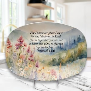 Puede incluir: Plato decorativo ovalado con un diseño de acuarela que presenta un campo de flores silvestres y un paisaje montañoso. El plato tiene el texto "For I know the plans I have for you...Jeremiah 29:11."