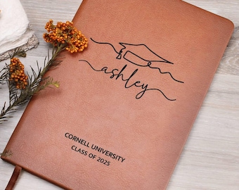 May include: A brown leather-bound journal with the name "ashley" in cursive and a graduation cap design. The journal is embossed with "Cornell University Class of 2025". A brown ribbon bookmark is visible.