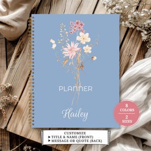 May include: A light blue spiral-bound planner featuring a watercolour floral bouquet illustration in pink, yellow, and white. The cover displays the text PLANNER and the name Hailey in white script. A pink circular badge notes 8 COLOURS 2 SIZES.