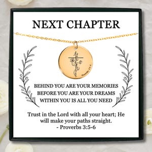 May include: A gold-colored necklace with a round pendant featuring a cross and floral design. The pendant hangs from a delicate chain. The card behind the necklace reads "NEXT CHAPTER" and includes inspirational text.