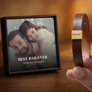 May include: A framed photo with the text "BEST DAD EVER" and a brown leather bracelet with a gold-colored clasp that reads "BEST DAD". The photo shows a man and a child. The bracelet is held by a hand.
