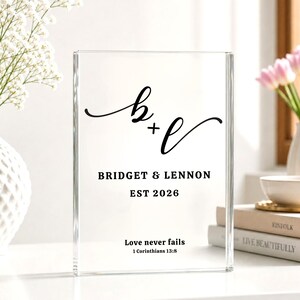 May include: Clear acrylic block with black script initials and the names Bridget & Lennon, established in 2026. The text "Love never fails" and a Bible verse are also printed on the block. A decorative wedding or anniversary keepsake.