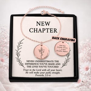 May include: Rose gold bracelet and pendant set with the words "NEW CHAPTER" and inspirational text. The pendant features a cross and floral design. The set is presented in a black box with floral accents.