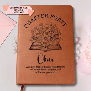 Peut inclure: Un carnet en similicuir beige avec le texte "CHAPTER FORTY" au-dessus d'un dessin au trait d'un livre ouvert avec un bouquet de fleurs. Sous l'illustration, le nom "Olivia" est imprimé, suivi du texte "As a new chapter begins, walk forward with confidence, purpose, and unlimited potential."