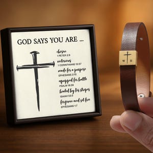 May include: A framed print with the text "GOD SAYS YOU ARE..." and a cross graphic, alongside a brown leather bracelet with a gold-colored square accent featuring a cross and initials.
