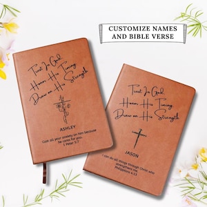 May include: Two tan leather-like journals featuring black engraved text. The left journal displays the name ASHLEY, a floral cross, and 1 Peter 5:7. The right journal displays the name JASON, a simple cross, and Philippians 4:13. Both share the text Trust In God. Honor His Timing. Draw on His Strength.