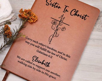 Op de afbeelding: Een beige notitieboek in lederlook met de tekst Sister In Christ, een bloemrijke kruisillustratie en het bijbelvers Galatians 6:2. De kaft bevat de naam Elisabeth en tekst over elkaar steunen in Zijn genade.