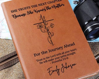 May include: A tan leather-like journal featuring a black engraved cross entwined with floral vines. The text reads: SHE TRUSTS THE NEXT CHAPTER Because She Knows the Author, For the Journey Ahead, Trust in the Lord with all your heart; He will make your paths straight. - Proverbs 3:5-6, and the name Emily Johnson.