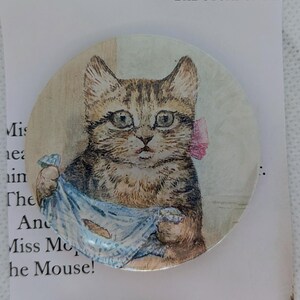 May include: A circular pin featuring a watercolor illustration of a brown tabby kitten with a pink bow behind its ear. The kitten is holding a blue and white patterned cloth. The background includes partial text from a storybook page.
