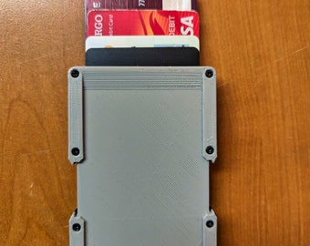 May include: A grey 3D printed card holder with a black card inside. The card holder has a textured surface and a red accent on the bottom right corner. The card holder is on a wooden surface.