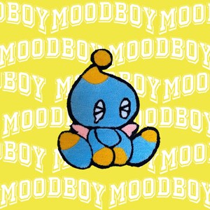 Könnte beinhalten: Ein blaues und gelbes Plüschtier mit einem traurigen Gesichtsausdruck. Das Spielzeug hat einen gelben Hut und rosa Flügel. Das Spielzeug sitzt auf einem gelben Hintergrund mit dem Text "MOODBOY" wiederholt.