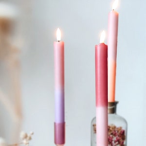 Op de afbeelding: Drie roze, ombre-kaarsen branden en staan in glazen kaarsenhouders op een houten tafel. De kaarsen zijn in verschillende tinten roze en wit. De kaarsenhouders zijn van helder glas en hebben een decoratief ontwerp.