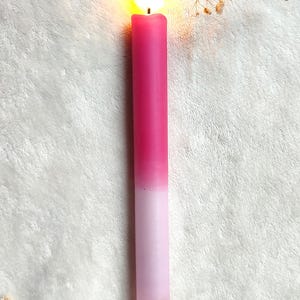 May include: A pink and white ombre candle with a flame burning at the top. The candle is sitting on a white fuzzy surface.