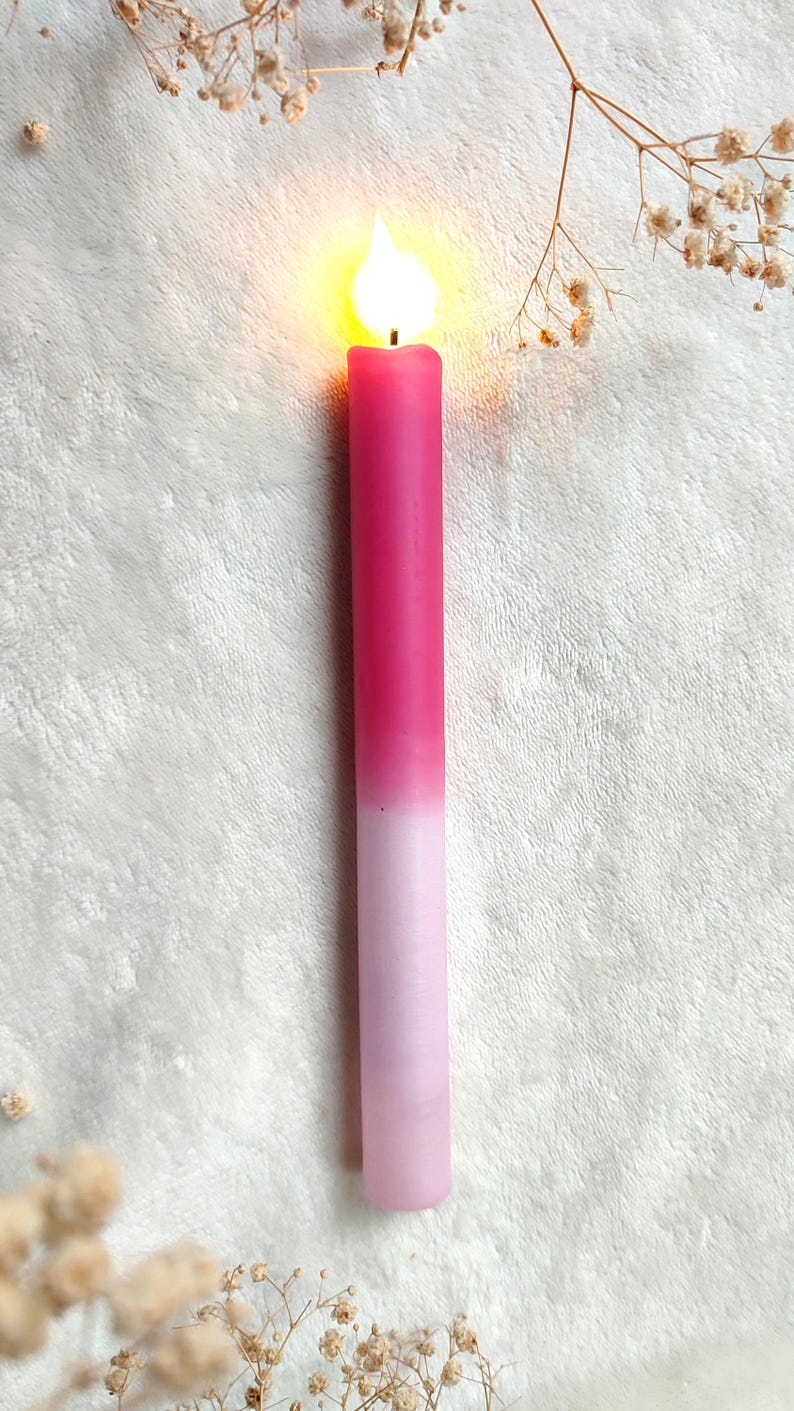 May include: A pink and white ombre candle with a flame burning at the top. The candle is sitting on a white fuzzy surface.