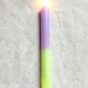 May include: A purple and green candle with a flame burning at the top. The candle is on a white fluffy surface.