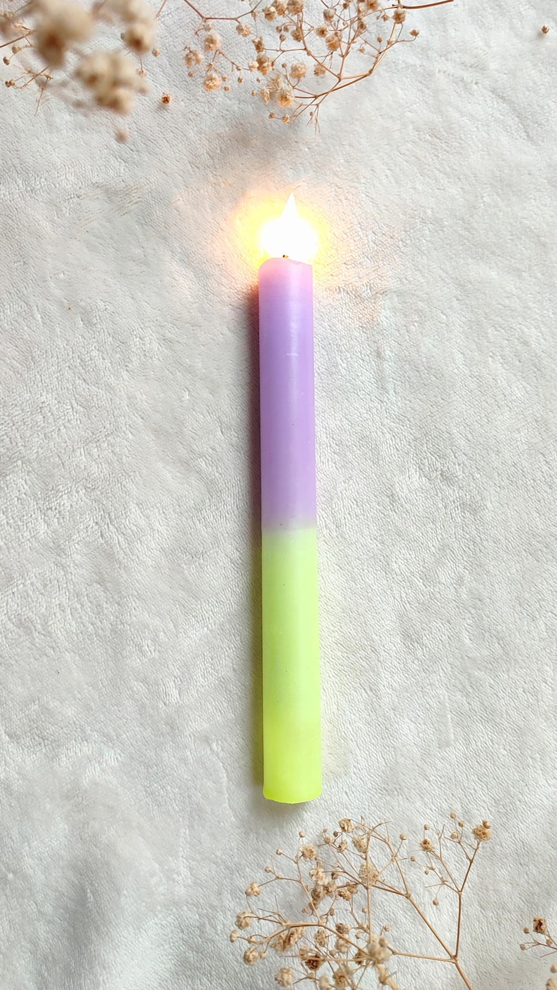 May include: A purple and green candle with a flame burning at the top. The candle is on a white fluffy surface.