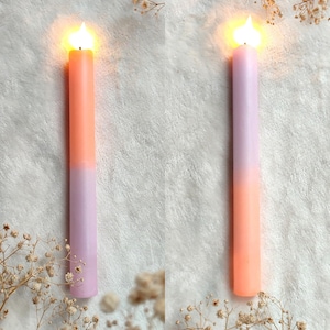 May include: Two lit candles with a gradient of peach and lavender colors. The candles are on a white fluffy surface.