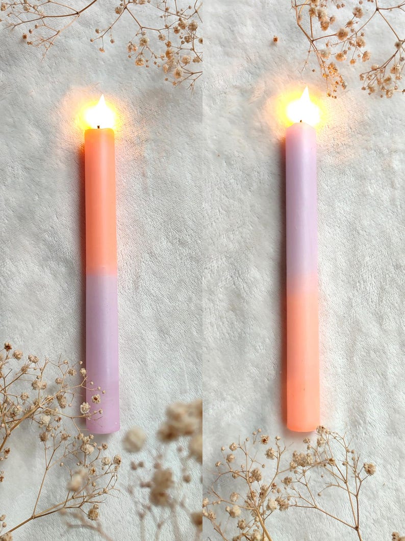 May include: Two lit candles with a gradient of peach and lavender colors. The candles are on a white fluffy surface.