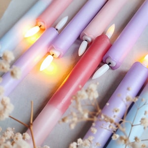 May include: Assortment of ombre taper candles in shades of pink, purple, and blue. The candles are arranged in a diagonal pattern and are lit, casting a warm glow.