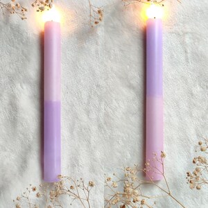 May include: Two lit candles with a light purple and pink ombre design. The candles are on a white fluffy surface with dried white flowers.