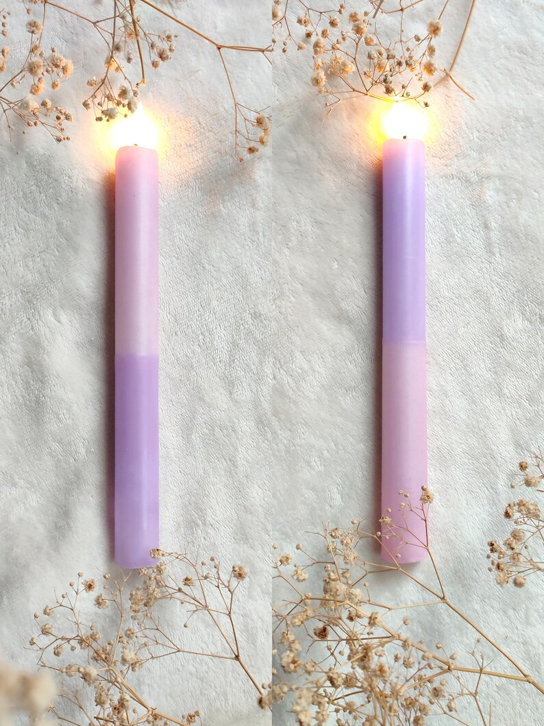May include: Two lit candles with a light purple and pink ombre design. The candles are on a white fluffy surface with dried white flowers.