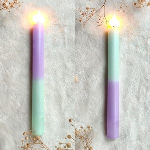 May include: Two lit candles with a gradient of purple and light green. The candles are on a white surface with dried flowers.