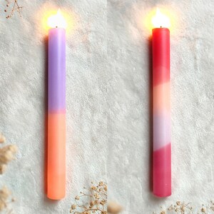 May include: Two lit candles with a gradient of colors. The candle on the left is purple and orange. The candle on the right is red, pink, and orange.