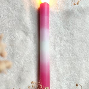 May include: A pink and white ombre candle with a lit flame on a white fuzzy surface.