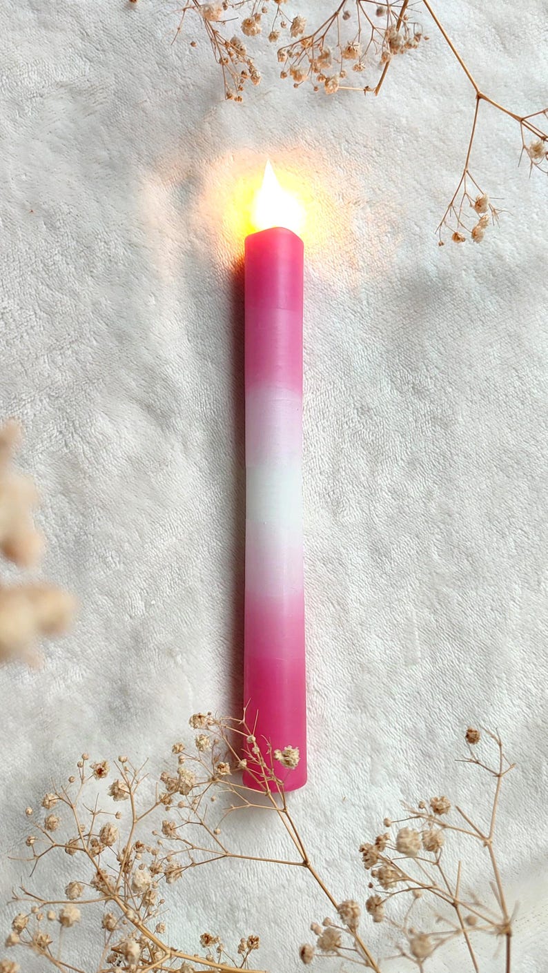 May include: A pink and white ombre candle with a lit flame on a white fuzzy surface.