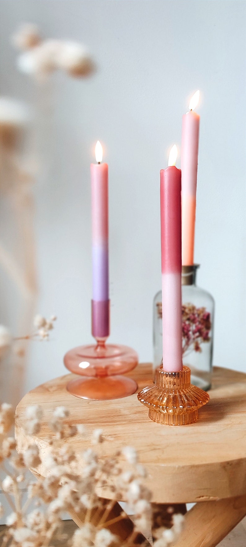 May include: Three pink, ombre candles are lit and placed in glass candle holders on a wooden table. The candles are in different shades of pink and white. The candle holders are clear glass and have a decorative design.