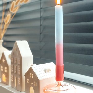 May include: A pink and blue ombre candle with a flame burning in a clear glass candle holder with a pink base. The candle holder is sitting on a white surface with white ceramic houses in the background.