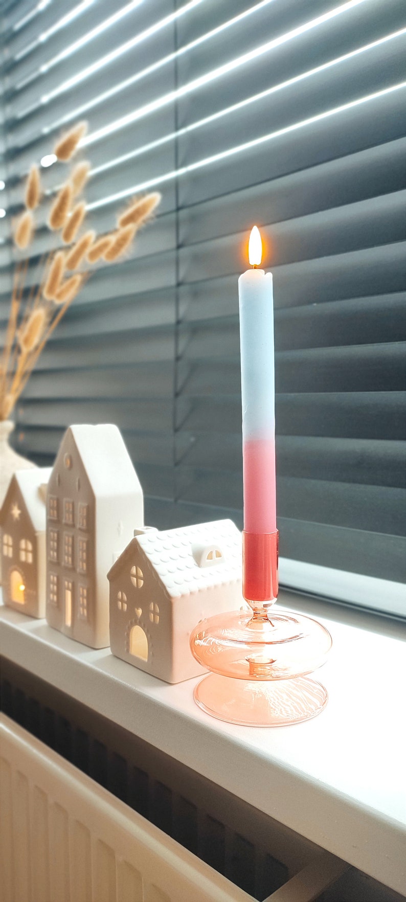 May include: A pink and blue ombre candle with a flame burning in a clear glass candle holder with a pink base. The candle holder is sitting on a white surface with white ceramic houses in the background.