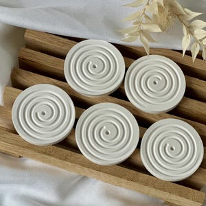 Könnte beinhalten: Fünf weiße, kreisförmige Untersetzer mit einem Spiraldesign sind auf einem Holztablett angeordnet. Die Untersetzer bestehen aus einem festen Material und haben ein kleines Loch in der Mitte. Das Tablett ist aus hellbraunem Holz.