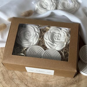 Könnte beinhalten: Eine Geschenkbox aus braunem Karton mit einem transparenten Fenster enthält weiße, rosenförmige und spiralförmige Ornamente. Die Box steht auf einer Holzoberfläche. Zwei Rosenornamente sind außerhalb der Box sichtbar.